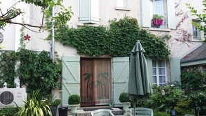 Une cour avec une table ronde en verre et des chaises, une porte en bois avec des volets et un pot de fleurs.