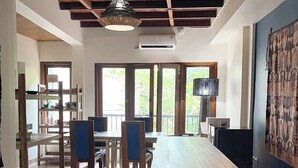Amplia habitación con techo alto, vigas de madera, una larga mesa y sillas. Ventanas que dejan entrar luz natural y ofrecen vistas a la vegetación exterior.