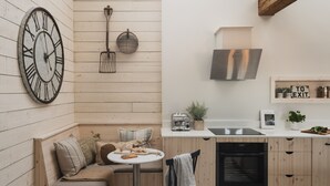 A modern kitchen with wooden cabinetry, a built-in oven, and a sink. There is a round table with a chair, a clock on the wall, and a shelf with kitchen items.