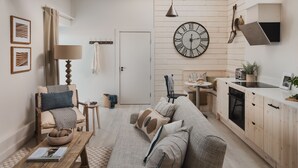 A cozy living room with a sofa, a coffee table, and a chair. There is a kitchen area with a stove, sink, and cabinets. A large clock is mounted on the wall.