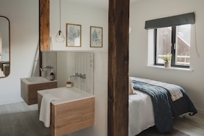 A bedroom with a bed, a window with a view, a wooden beam, and a bathroom with a sink and mirror.