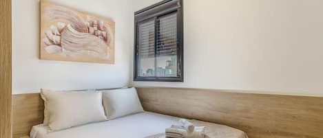 Una cama bien tendida con una colcha color crema, una almohada y una toalla doblada sobre un cabecero de madera. Un cuadro cuelga en la pared encima de la cama.