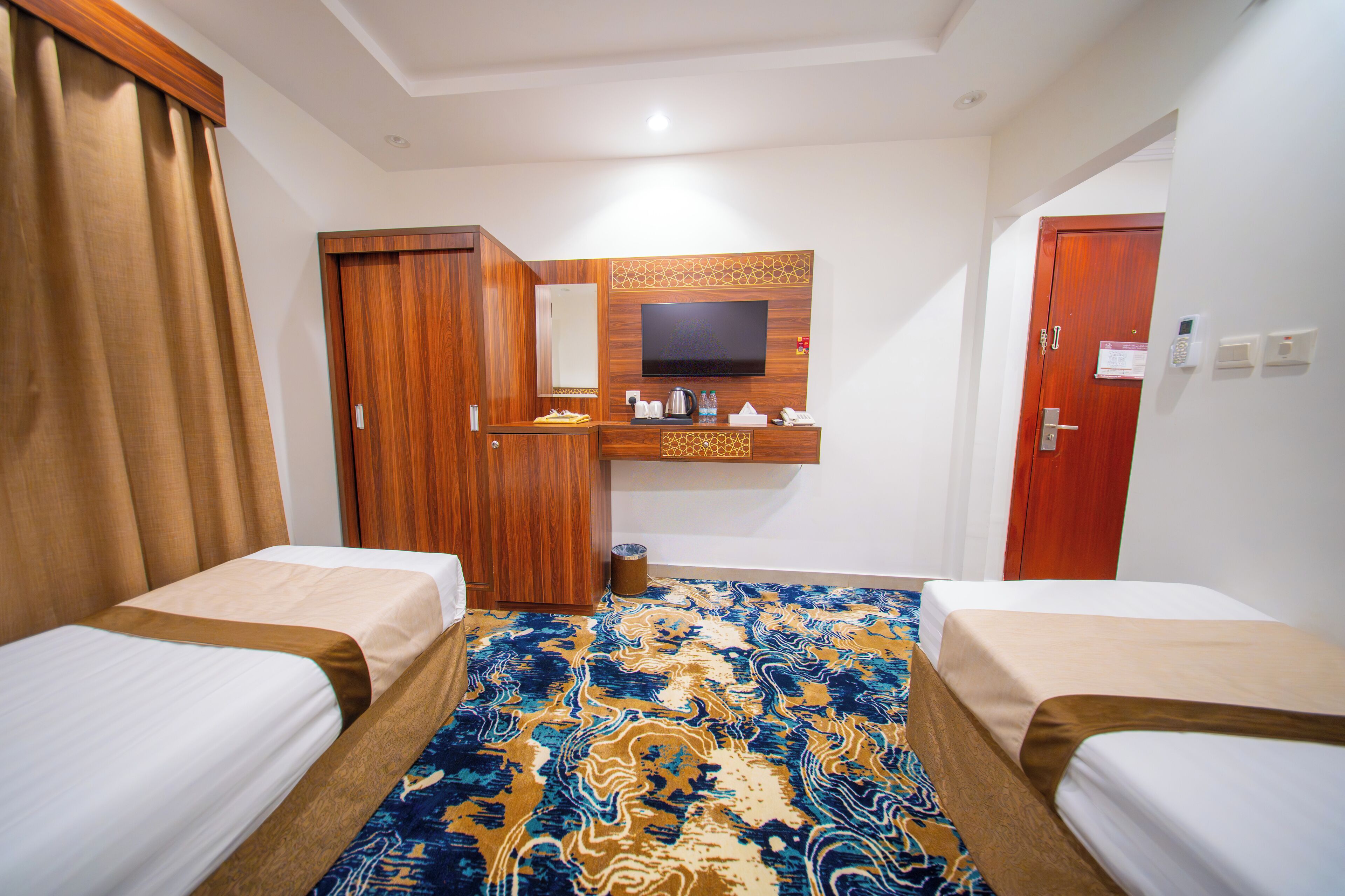 A hotel room with two beds, a television mounted on the wall, a wooden wardrobe, and a patterned carpet.