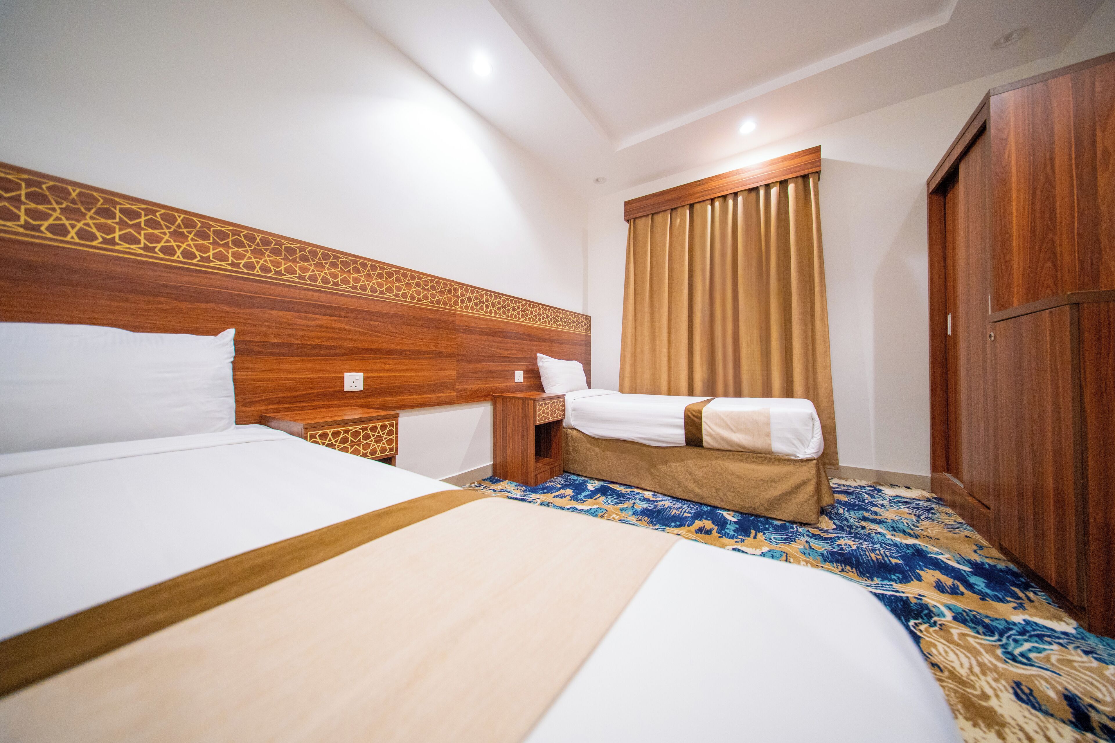 Habitación de hotel con dos camas, cabecero de madera, mesita de noche, armario y alfombra estampada.