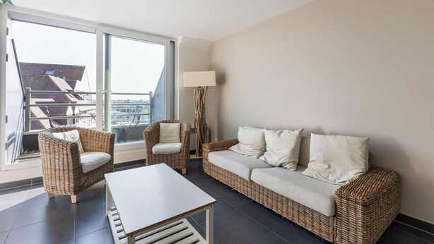 Una sala de estar moderna con sofá, dos sillones, una mesa de centro y una lámpara de pie. Amplias ventanas ofrecen vista a la ciudad.