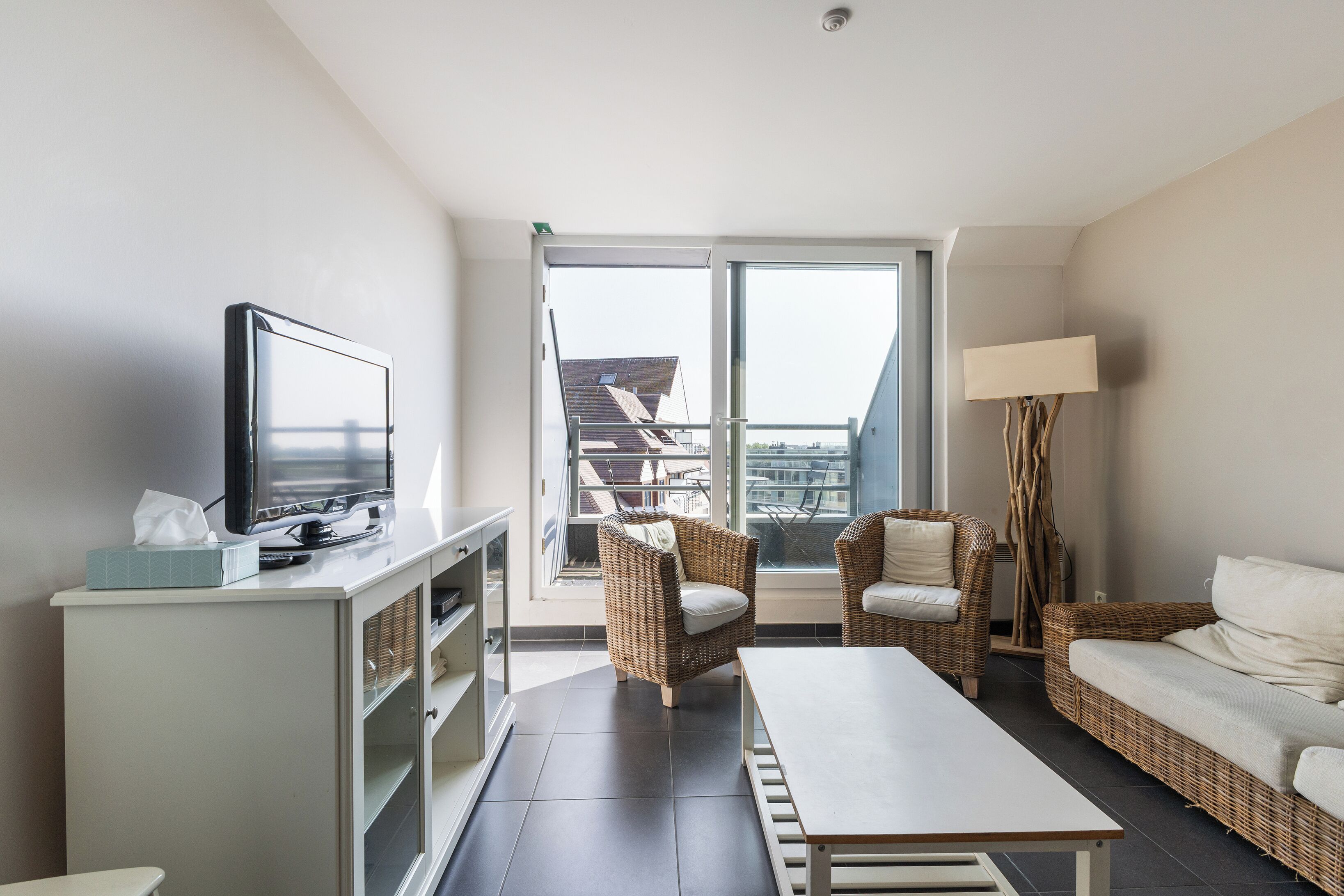 A modern living room with a television, wicker furniture, a sofa, a table, and a view of the city through large windows.