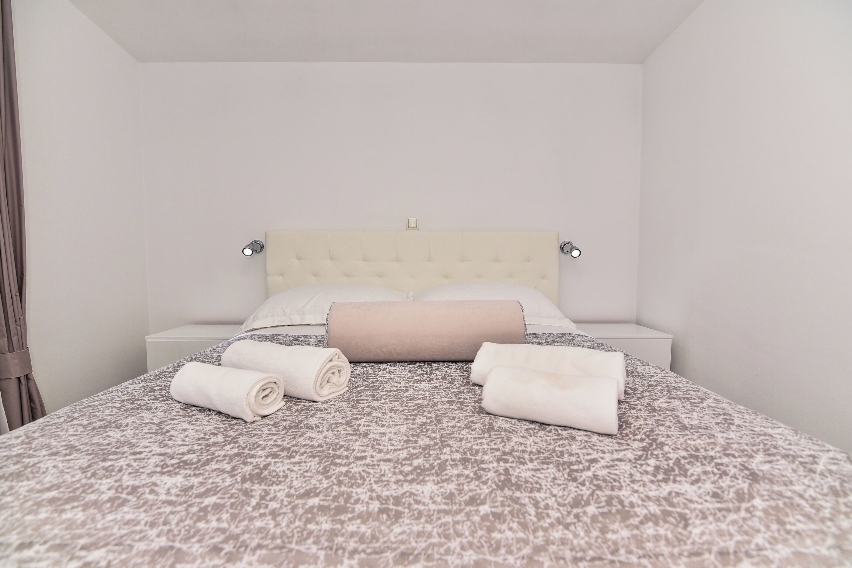 Un letto rifatto con cura, con copripiumino a fantasia, asciugamani arrotolati, una testiera imbottita e un comodino con una lampada.