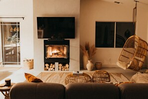 A cozy living room with a fireplace, a television mounted above it, and a hanging chair. - License To Chill | Pool + Hot Tub | Enclosed Yard (Yucca Valley)