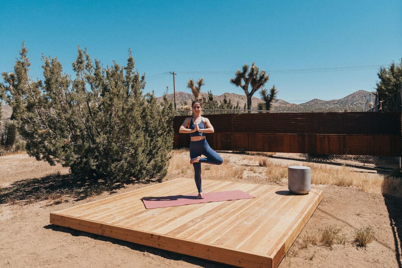 A person performing a yoga pose on a wooden platform outdoors with a clear sky and desert vegetation in the background.