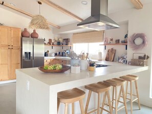 A modern kitchen with a central island, wooden stools, and a hanging light fixture. - License To Chill | Pool + Hot Tub | Enclosed Yard (Yucca Valley)