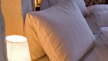 Una cama con ropa blanca y almohadas, una lámpara al lado y una pared de piedra al fondo.