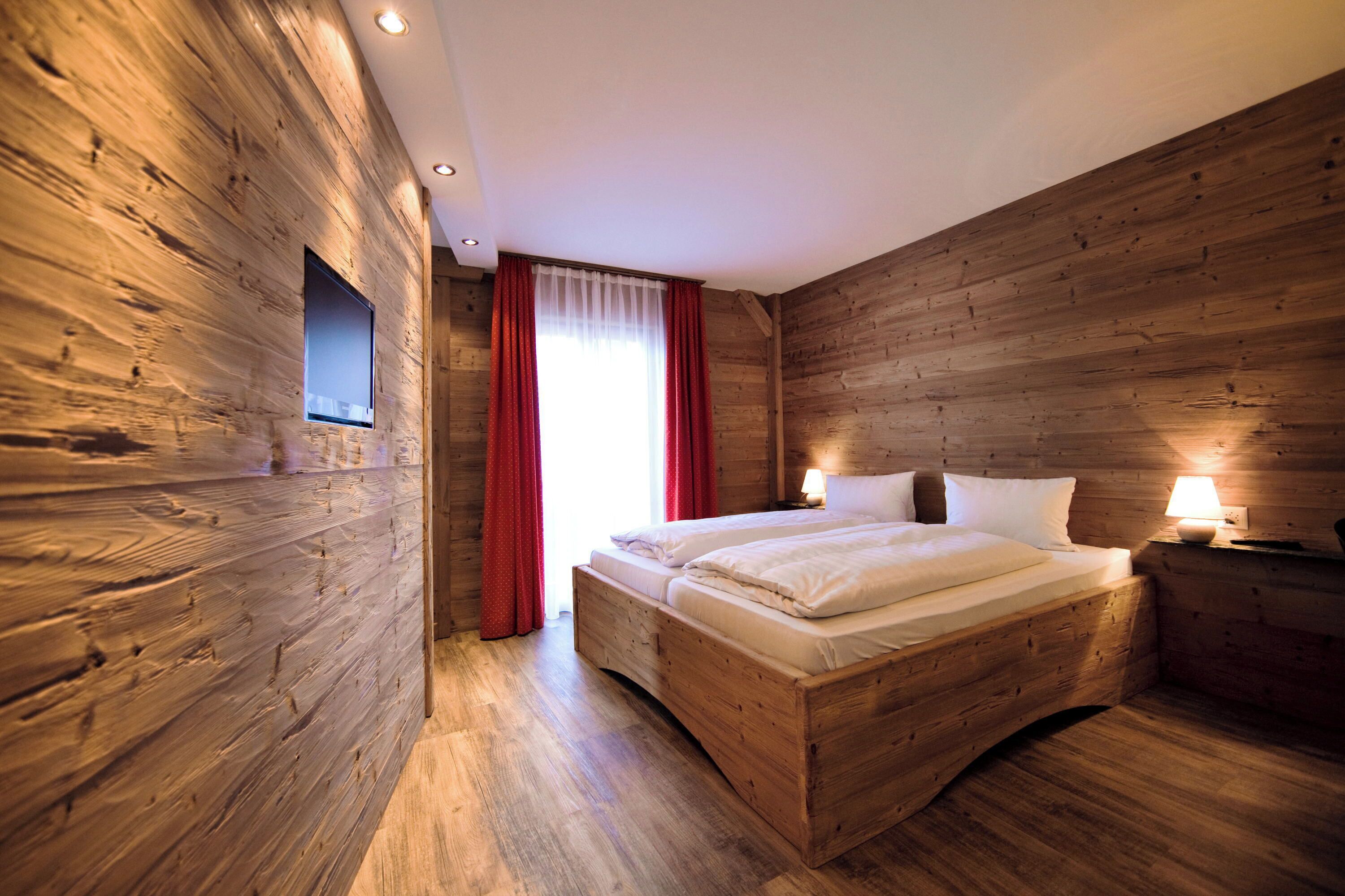 A wooden-paneled bedroom with a bed, two bedside lamps, and a window with red curtains.
