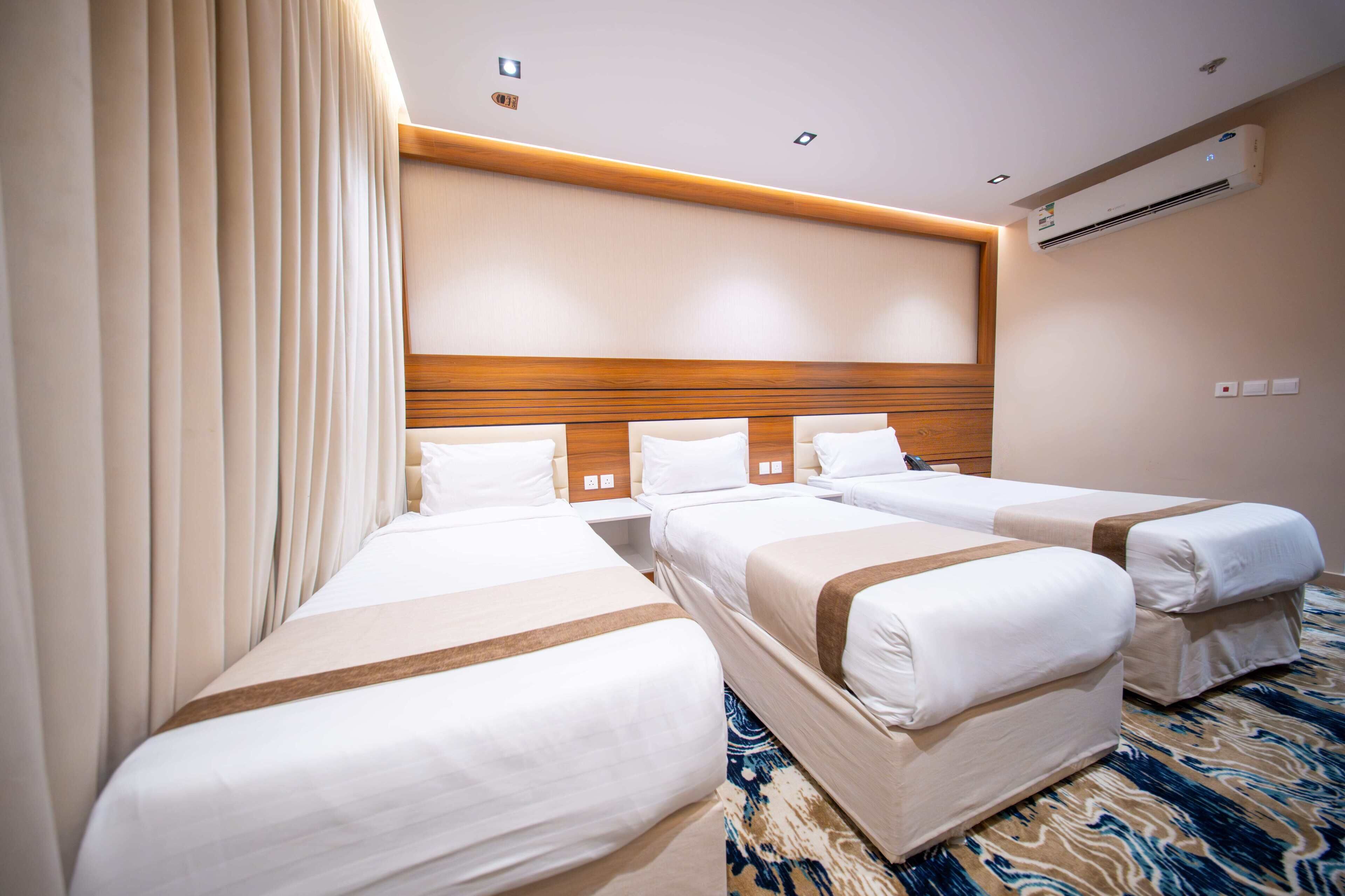 Habitación de hotel con tres camas individuales, cabecera de madera, cortinas color beige y alfombra estampada.