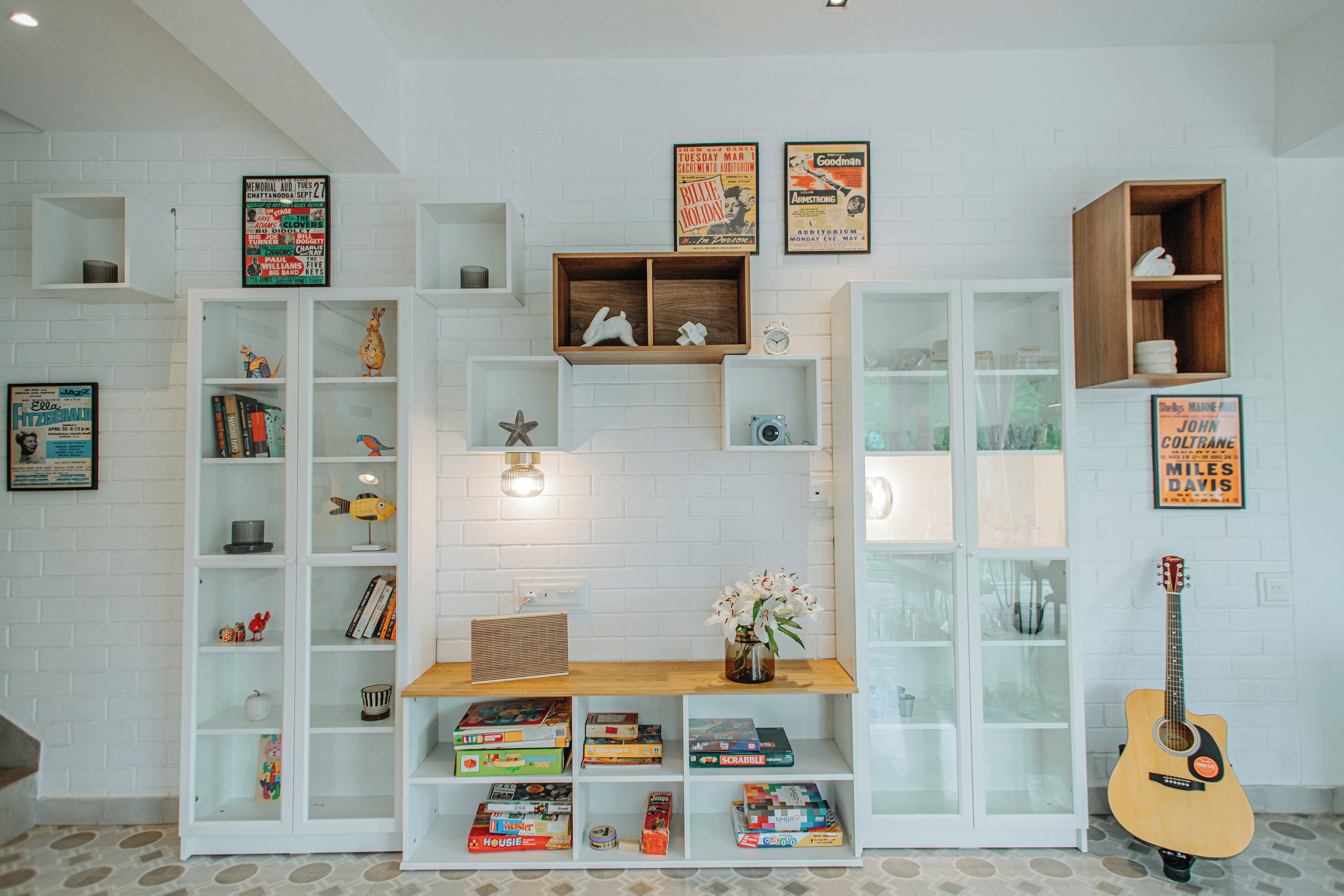 A room with white brick walls, multiple shelves with books and games, a guitar, and framed posters.