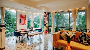 A modern living room with a dining area, large windows, and a view of greenery. - Palette by RÊVE/2BR Duplex/Art Getaway/Shared Pool (Assagao)