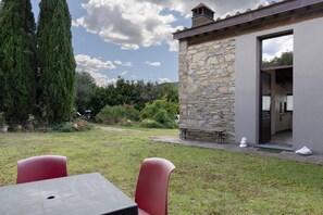 A stone building with a doorway leading to a kitchen area, surrounded by a grassy yard and tall trees.