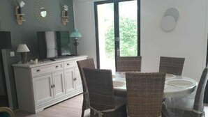 A dining area with wicker chairs and a white table, a white cabinet with a TV, and a window with a view of greenery.