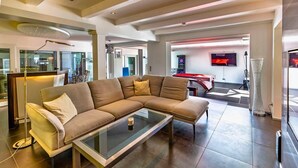 A modern living room with a sectional sofa, a glass coffee table, and a pool table in the background.
