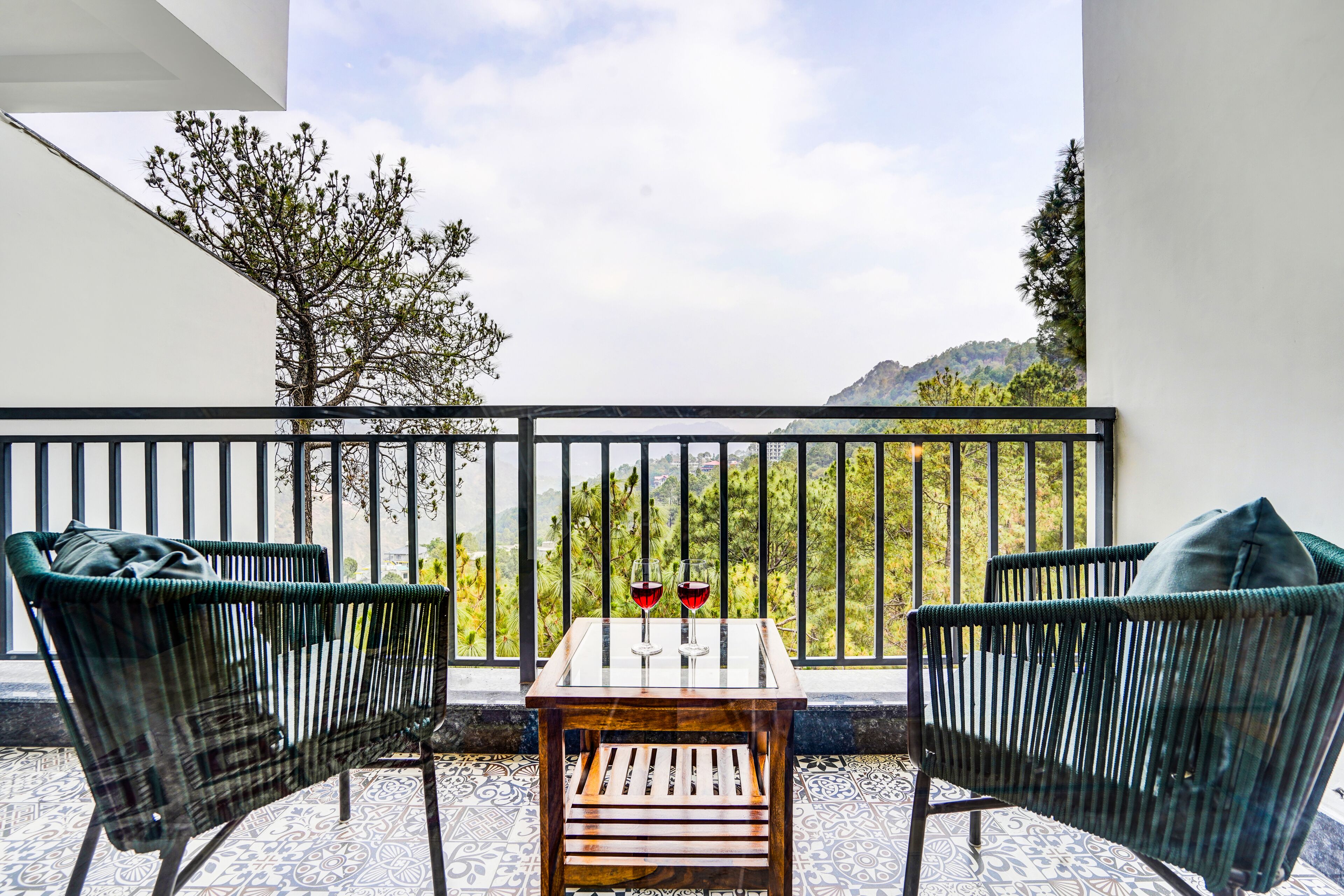 Un balcon avec une table et deux chaises, un verre de vin rouge et une vue sur un paysage montagneux.