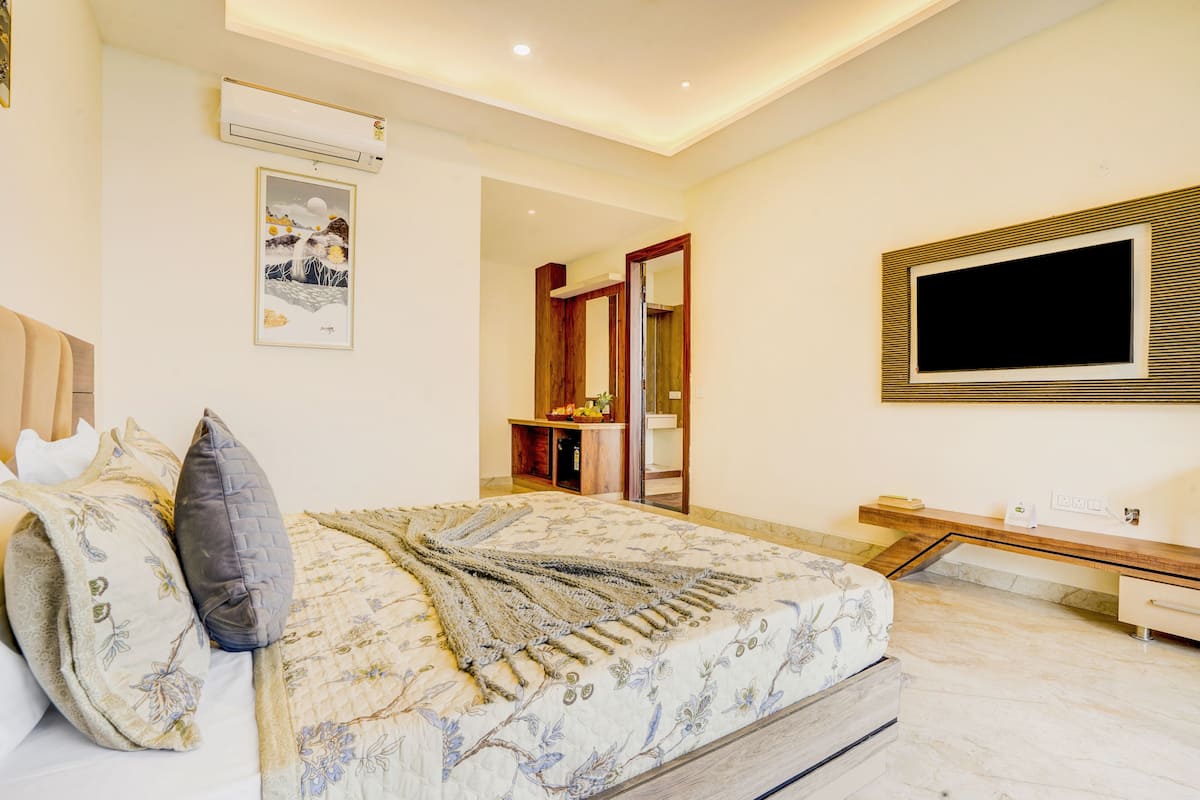 A bedroom with a bed, a TV mounted on the wall, a wooden bench, and a small table with a fruit bowl.