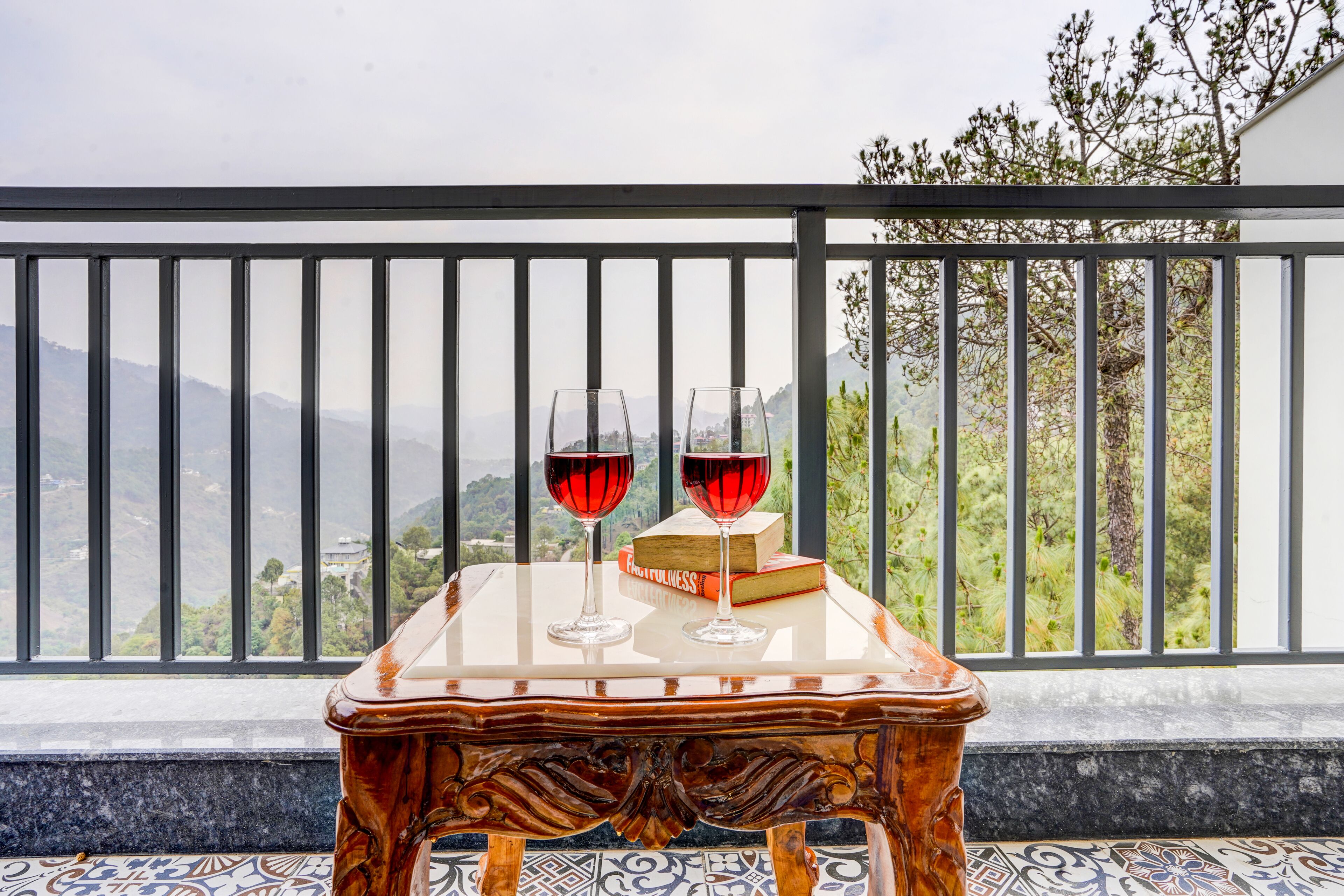 A table with two wine glasses and a book on a balcony with a view of mountains.