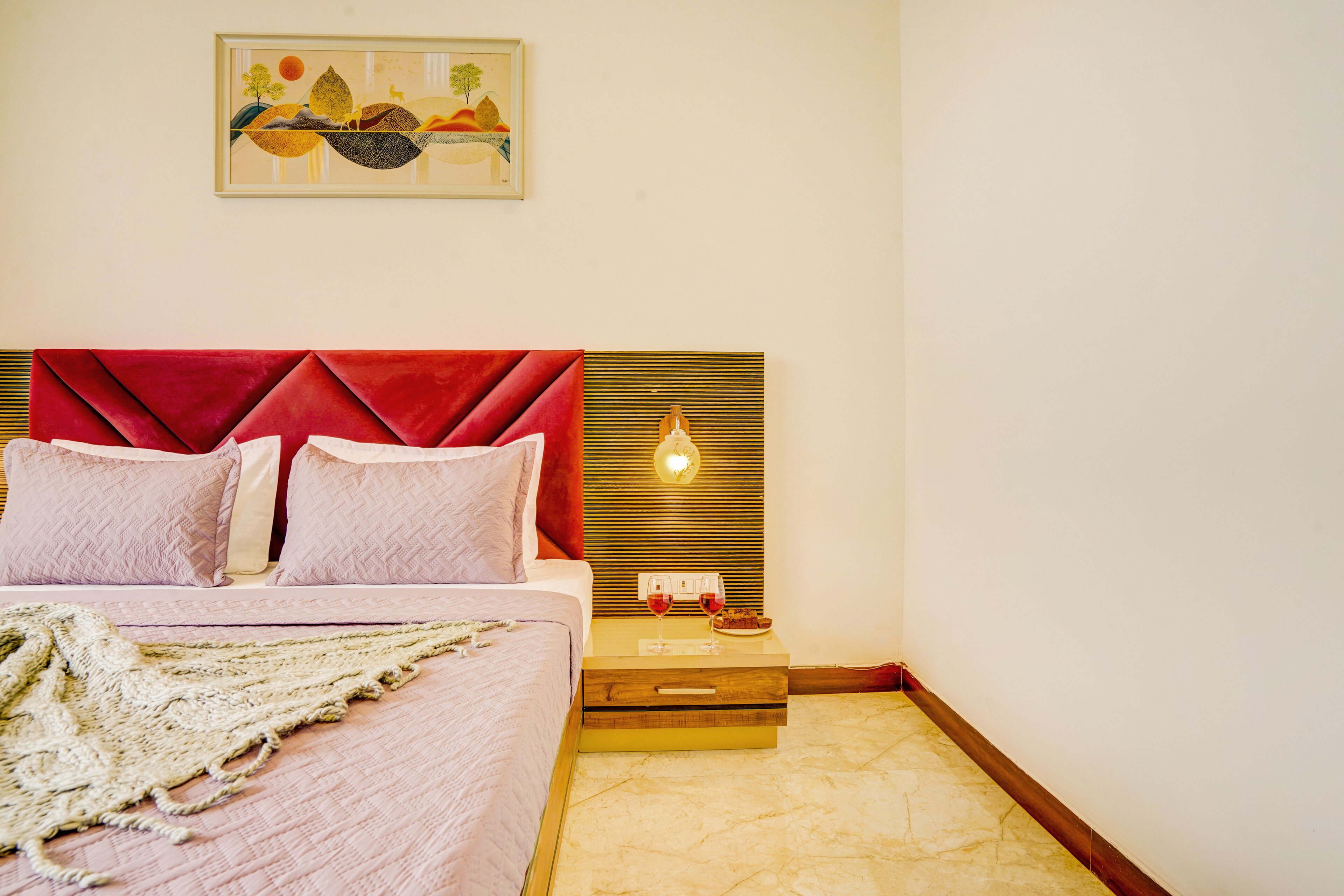 Una cama bien tendida con cabecera roja, una mesita de noche con lámpara y una obra de arte enmarcada sobre la cama.