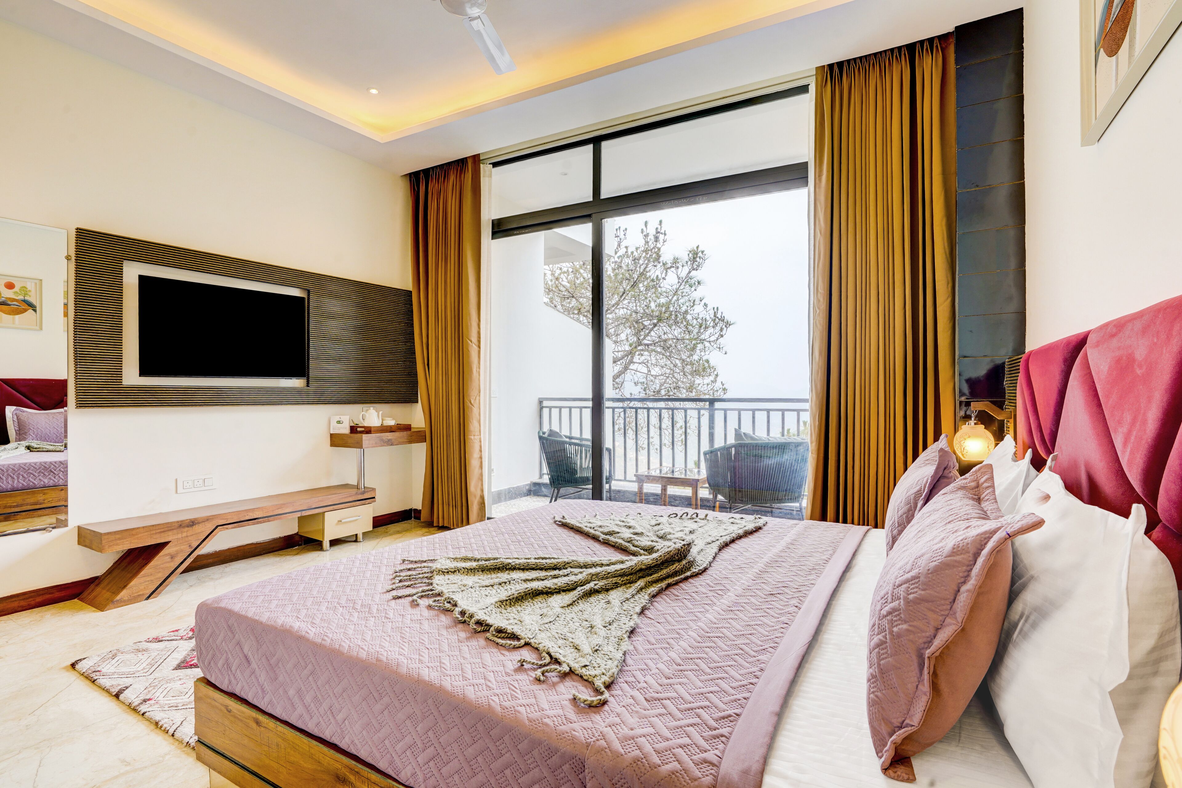 A modern bedroom with a large bed, a flat-screen TV mounted on the wall, a balcony with outdoor seating, and a large window with curtains.