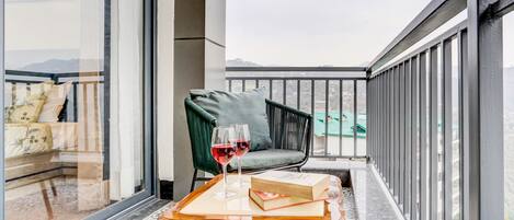 A balcony with a view of a cityscape, a green chair, a wooden table with a book and glasses of red wine.