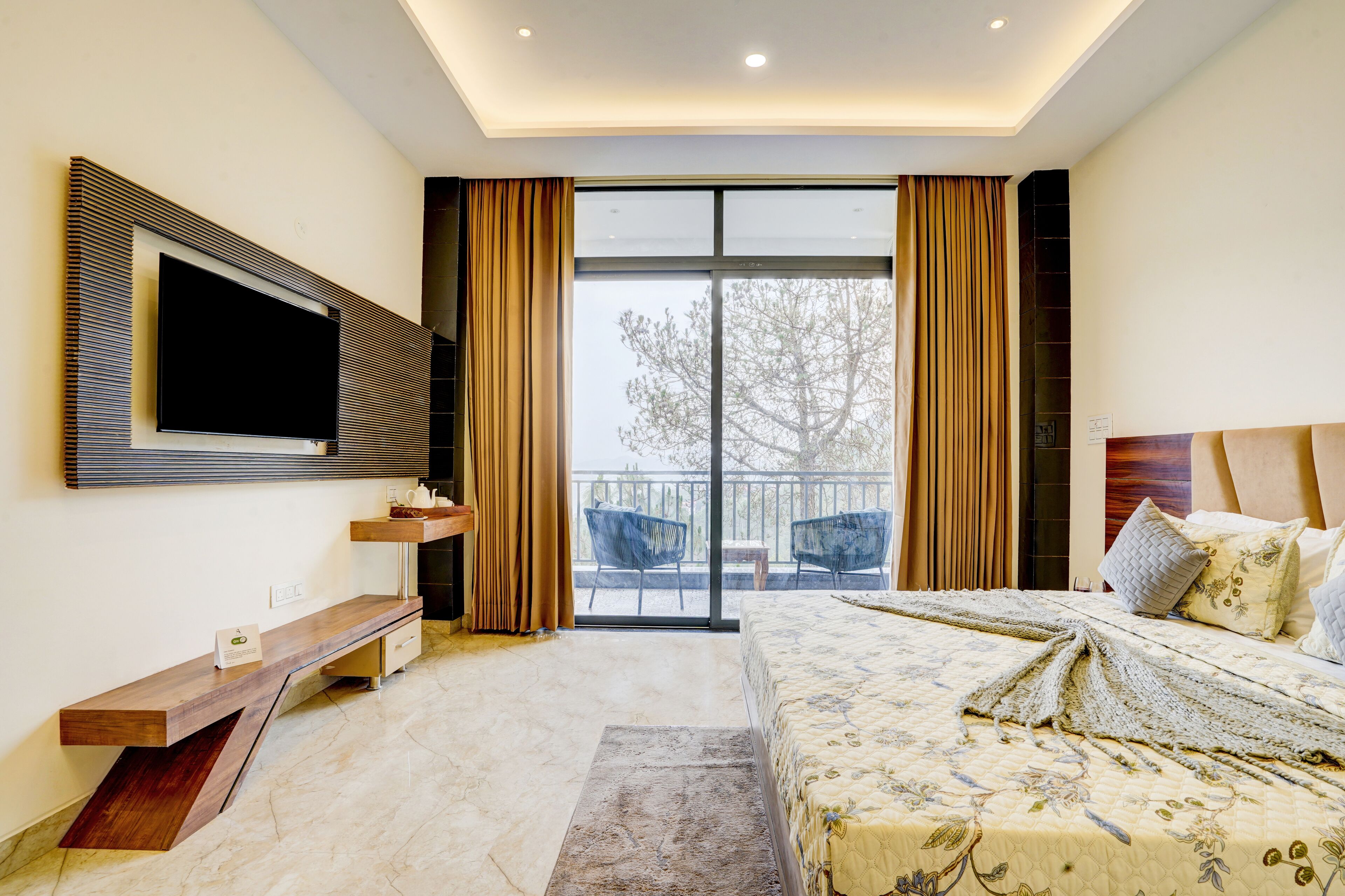 A modern hotel room with a large bed, a flat-screen TV mounted on the wall, a small desk with a chair, and a balcony with curtains.