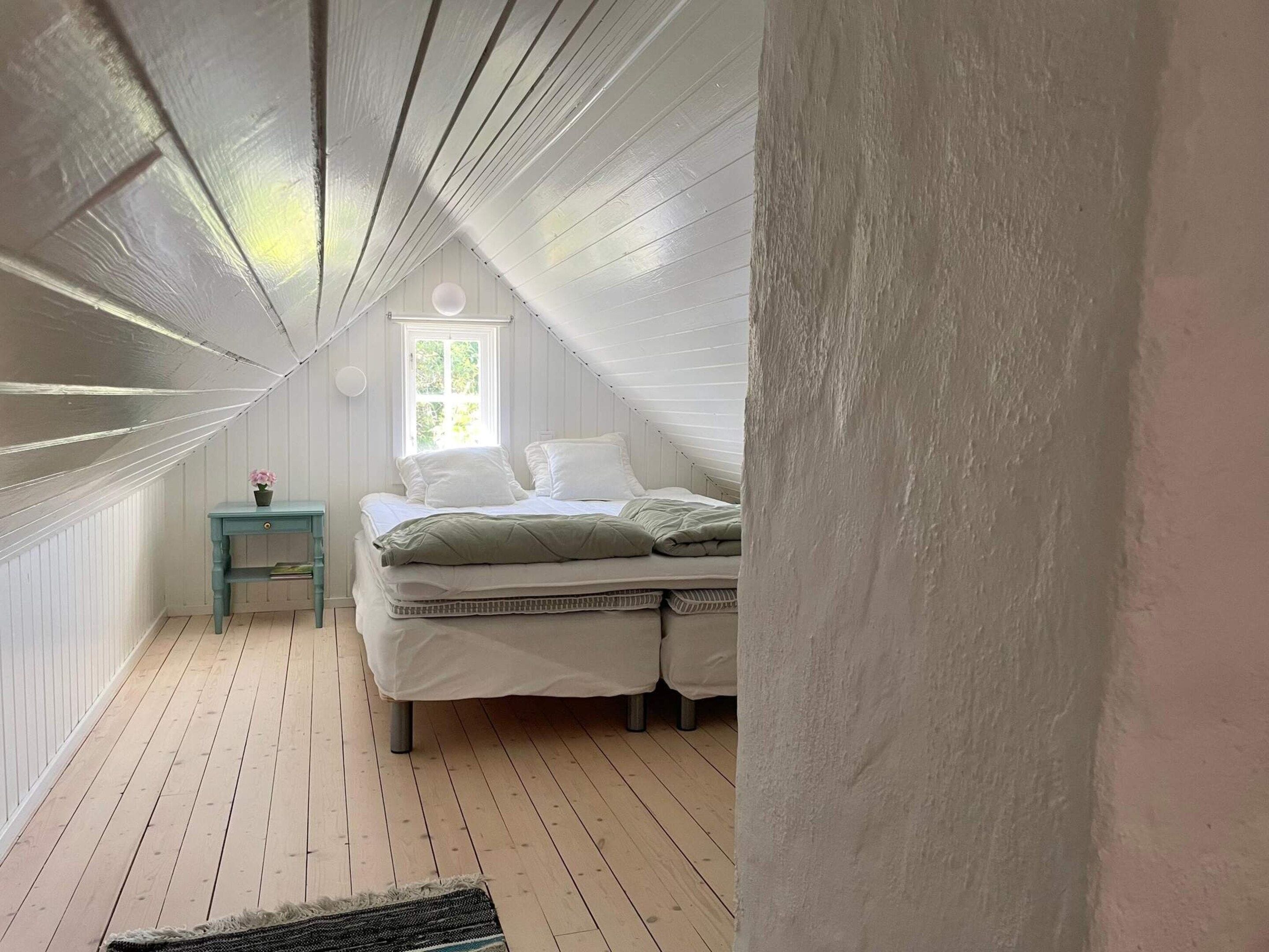 A bedroom with a sloped ceiling, a bed with white bedding, a small table with a flower, and a window with natural light.