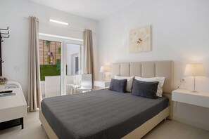 A modern hotel room with a large bed, two bedside lamps, a desk with a chair, and a view of a green outdoor area.