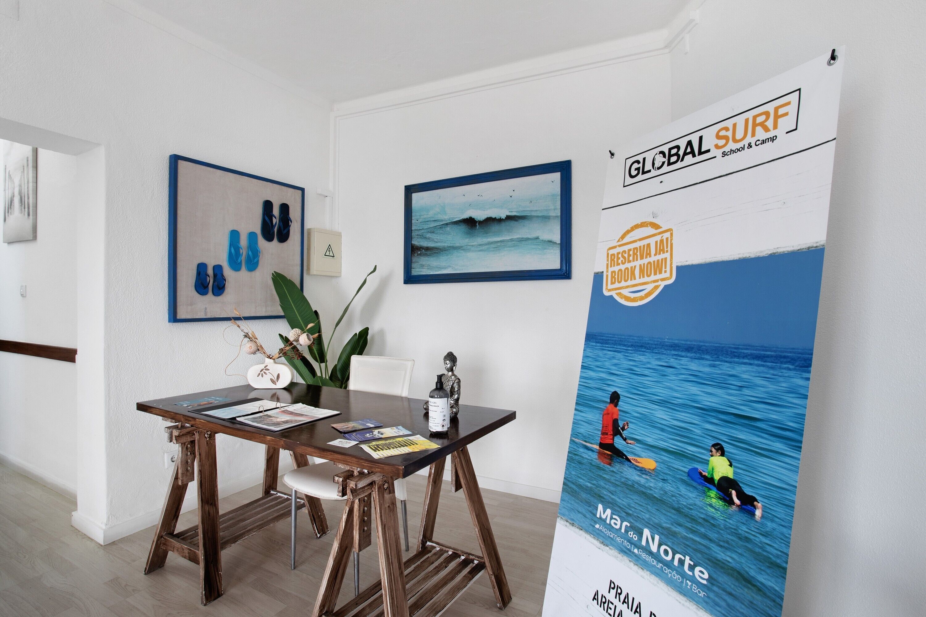 A room with a wooden table, framed artwork, and a poster advertising a surf school.