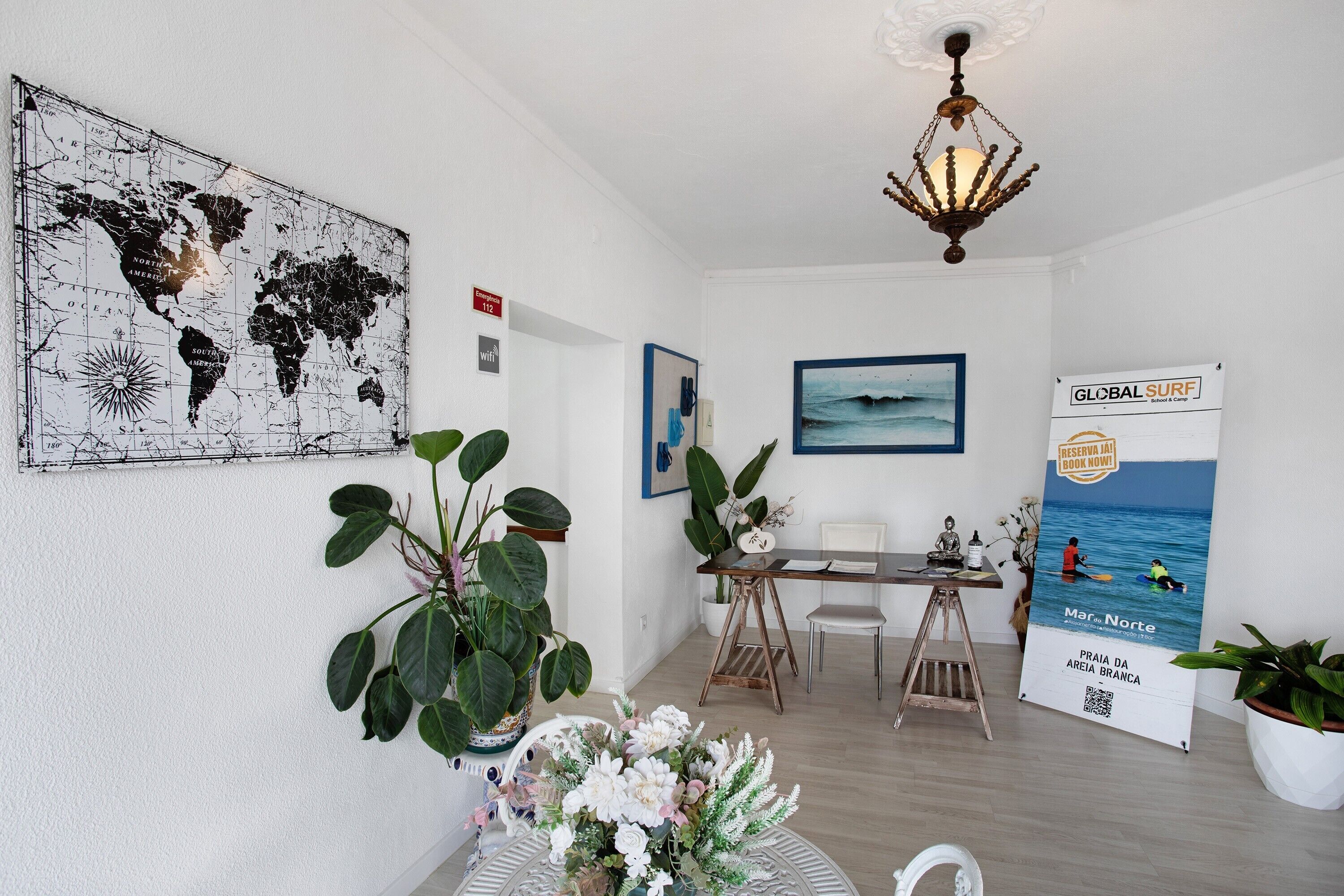 A room with a world map, a dining area with a table and chairs, a chandelier, and a poster advertising a surf event.