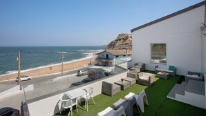 A balcony with a view of the beach and ocean, outdoor seating, and a building with a large window.