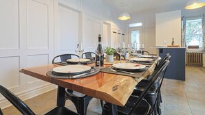 A dining area with a wooden table set for six, surrounded by black chairs. There are wine glasses, plates, and a bottle on the table. The room has white paneled walls and a window with curtains.