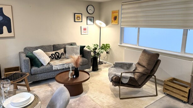 A modern living room with a grey sofa, a round coffee table, a brown armchair, and a large window with blinds. - Cozy Notting Hill 1 Bedroom - Prime Spot (London)