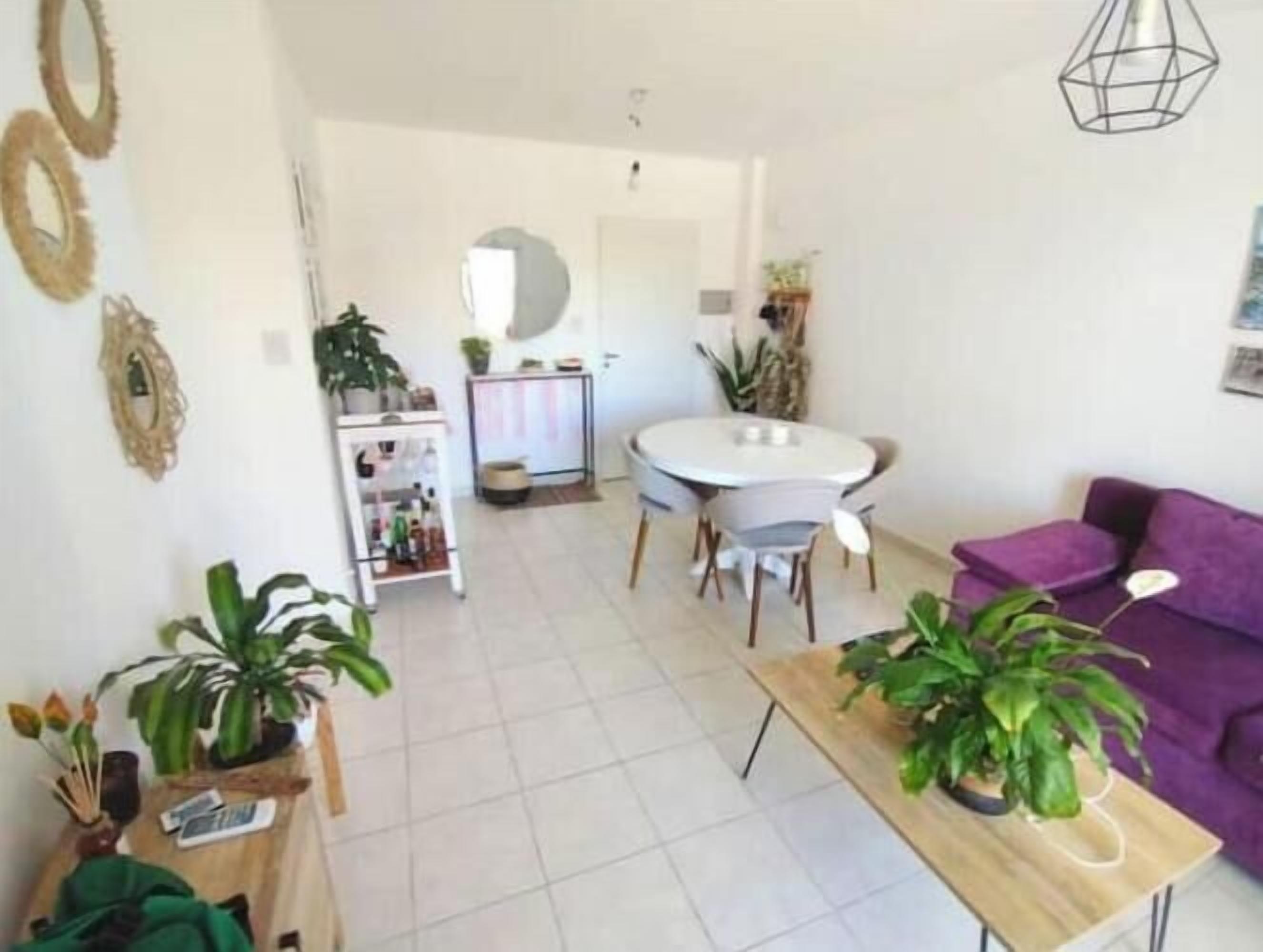 Un salon moderne avec un canapé violet, une table basse en bois et un coin repas comprenant une table et des chaises blanches. Plusieurs plantes en pot et un miroir rond ornent les murs.