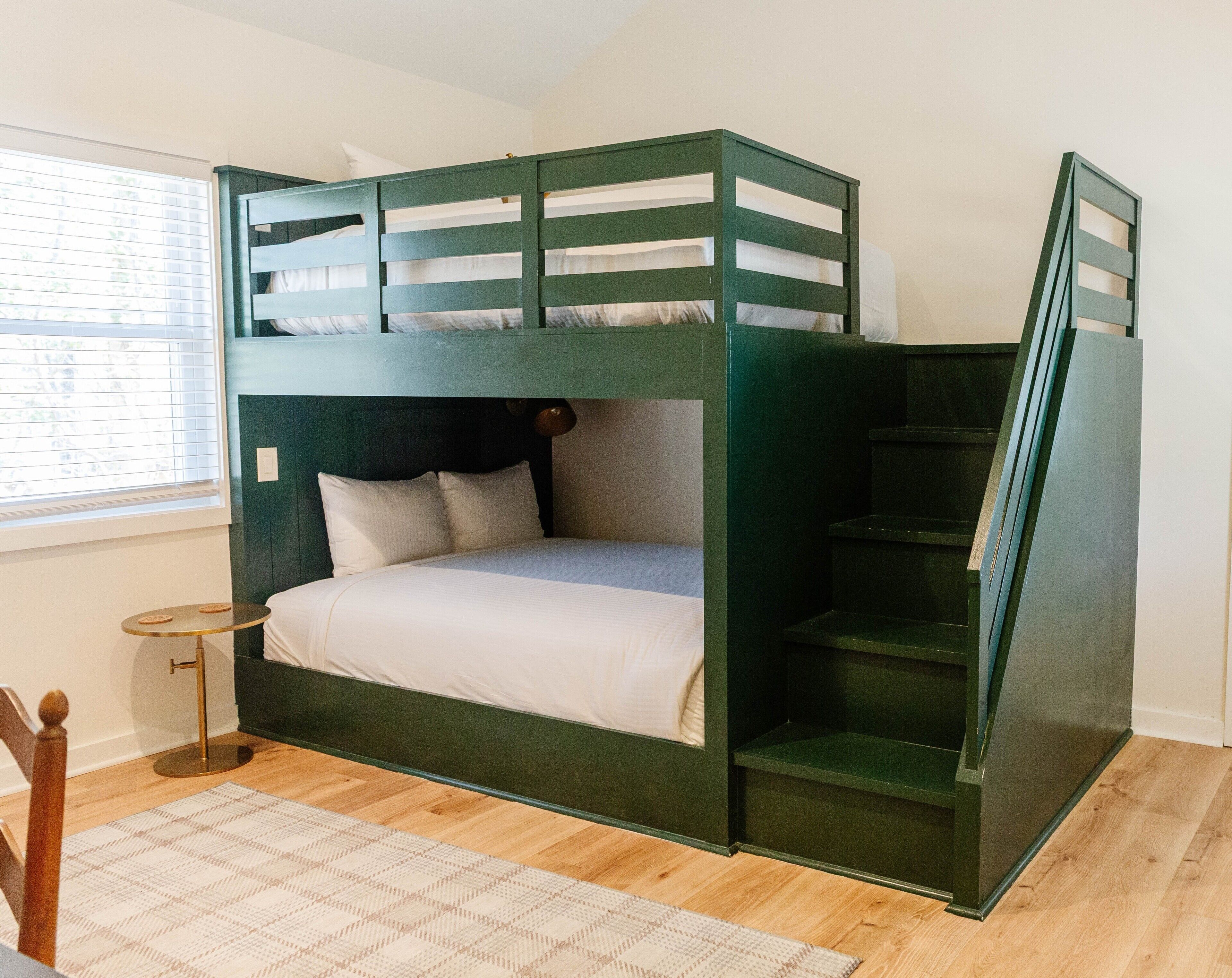 A bunk bed with a wooden frame, a small bedside table, and a window with blinds.