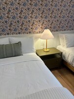 Una habitación de hotel con cama, mesita de noche con lámpara y papel tapiz estampado con motivos florales.