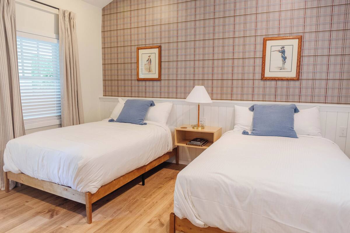 A hotel room with two single beds, a wooden floor, a plaid-patterned wall, and a window with curtains.