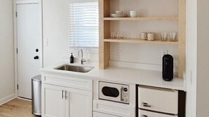 A compact kitchen with white cabinetry, a built-in microwave, a sink, and a shelf with glassware and a picture frame. - Rochambeau Roadside Inn (Williamsburg)
