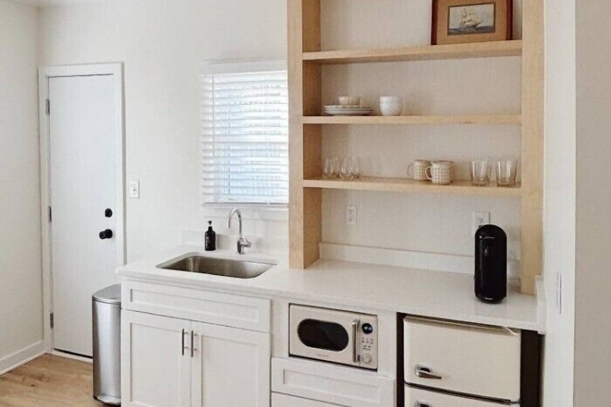 A compact kitchen with white cabinetry, a built-in microwave, a sink, and a shelf with glassware and a picture frame.