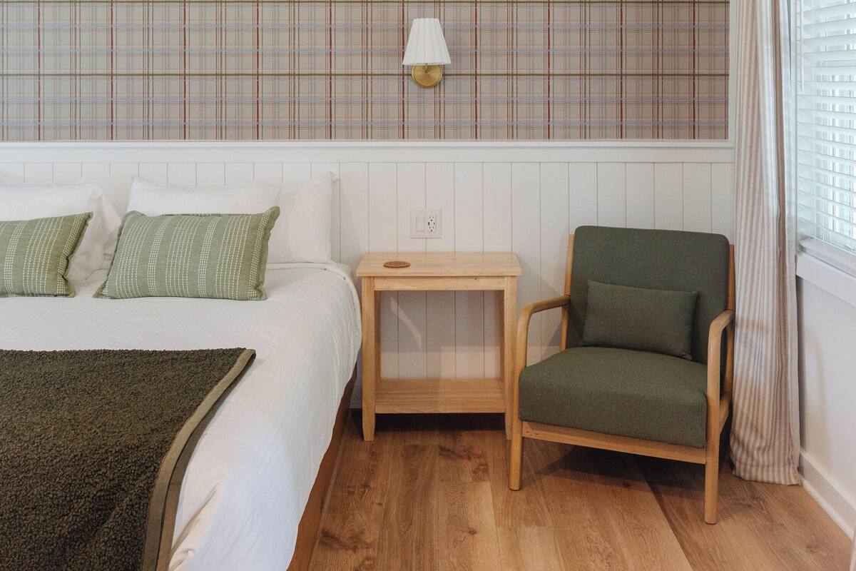 A bedroom with a plaid-patterned headboard, a wooden bedside table, a green armchair, and a window with curtains.