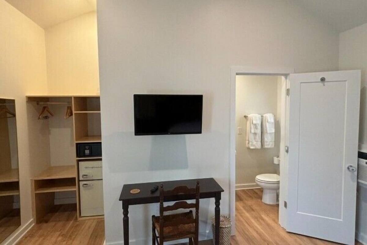 A room with a flat-screen TV mounted on the wall, a wooden table with a chair, a white door, and a bathroom visible through an open door.