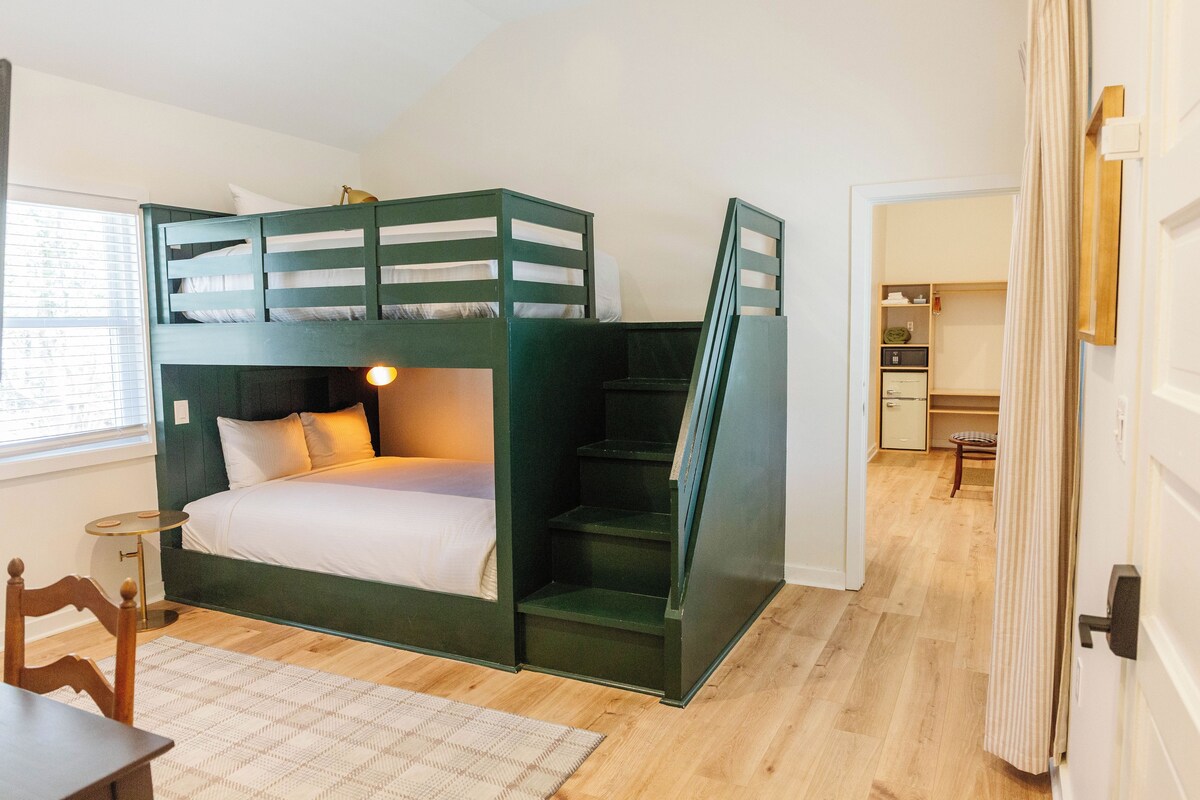 A bunk bed room with a desk and chair, a small table, and a window with blinds.