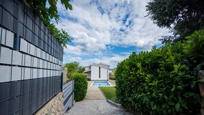 A pathway leading to a house with a swimming pool, surrounded by greenery and a stone wall.