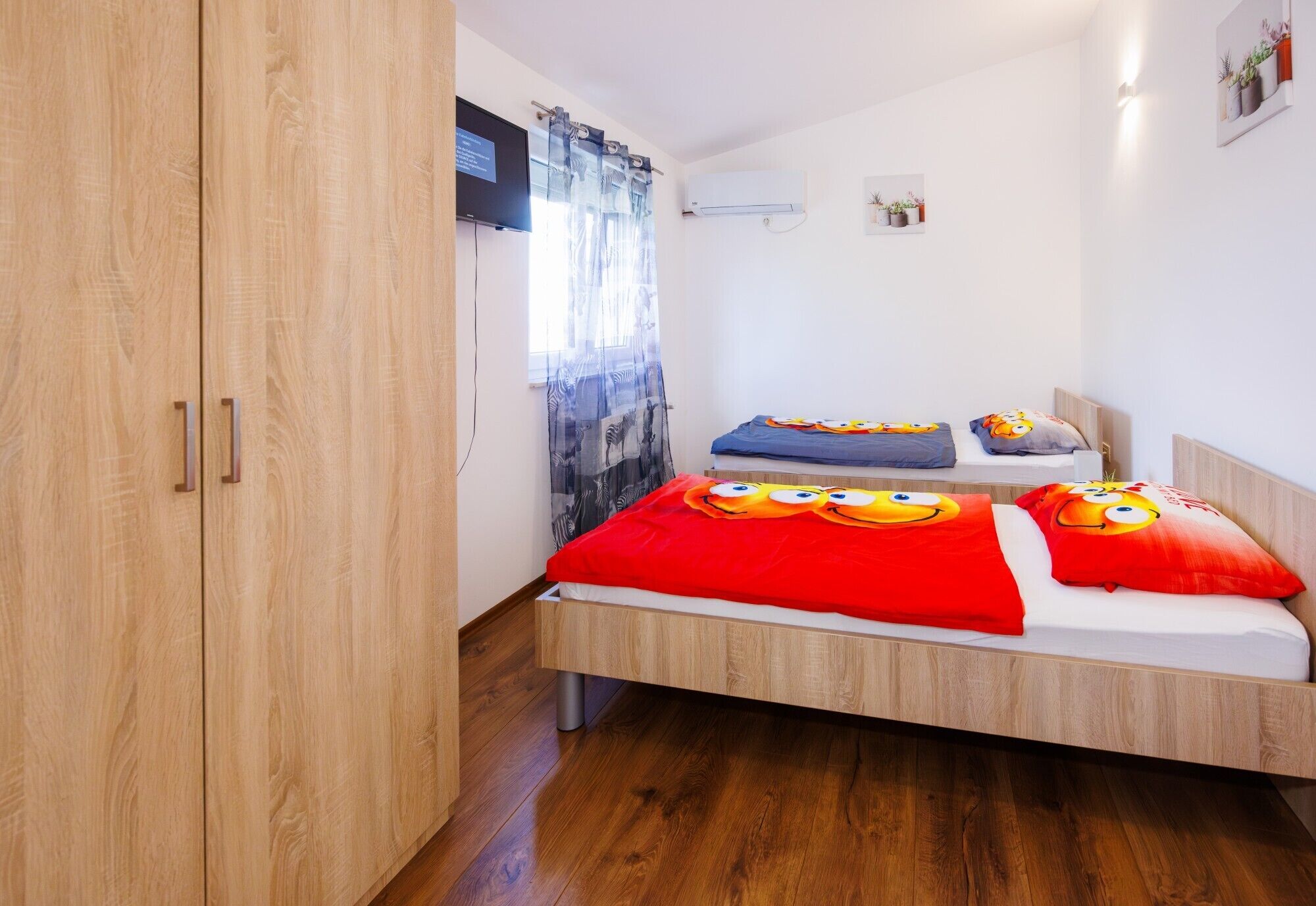 Un dormitorio pequeño y moderno con un armario de madera, una cama con una manta roja y almohadas con motivos de dibujos animados, y una ventana con cortinas estampadas.