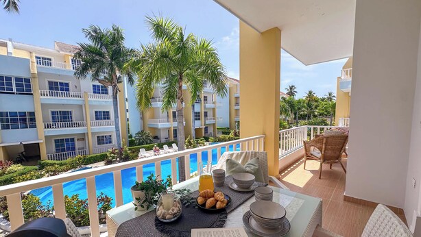 A balcony with a table set for breakfast, overlooking a swimming pool and adjacent buildings.