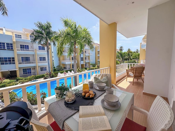 Un balcón con una mesa dispuesta para el desayuno, con vista a una piscina y edificios cercanos.