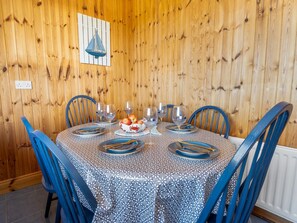 A wooden-paneled dining room with a round table set for six, blue chairs, and a decorative bowl of fruit.